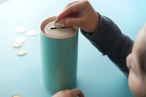 Anak kecil memasukkan uang koin ke dalam celengan transparan untuk belajar menabung